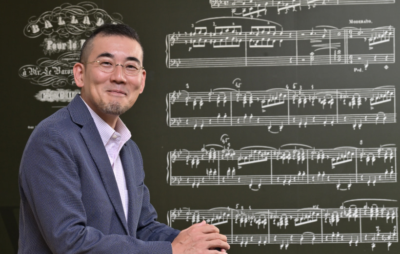 譜面を書いた黒板の前で、笑顔の足本准教授