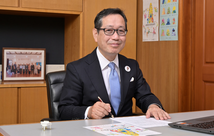 机で書き物しつつ、笑顔の梅本学長